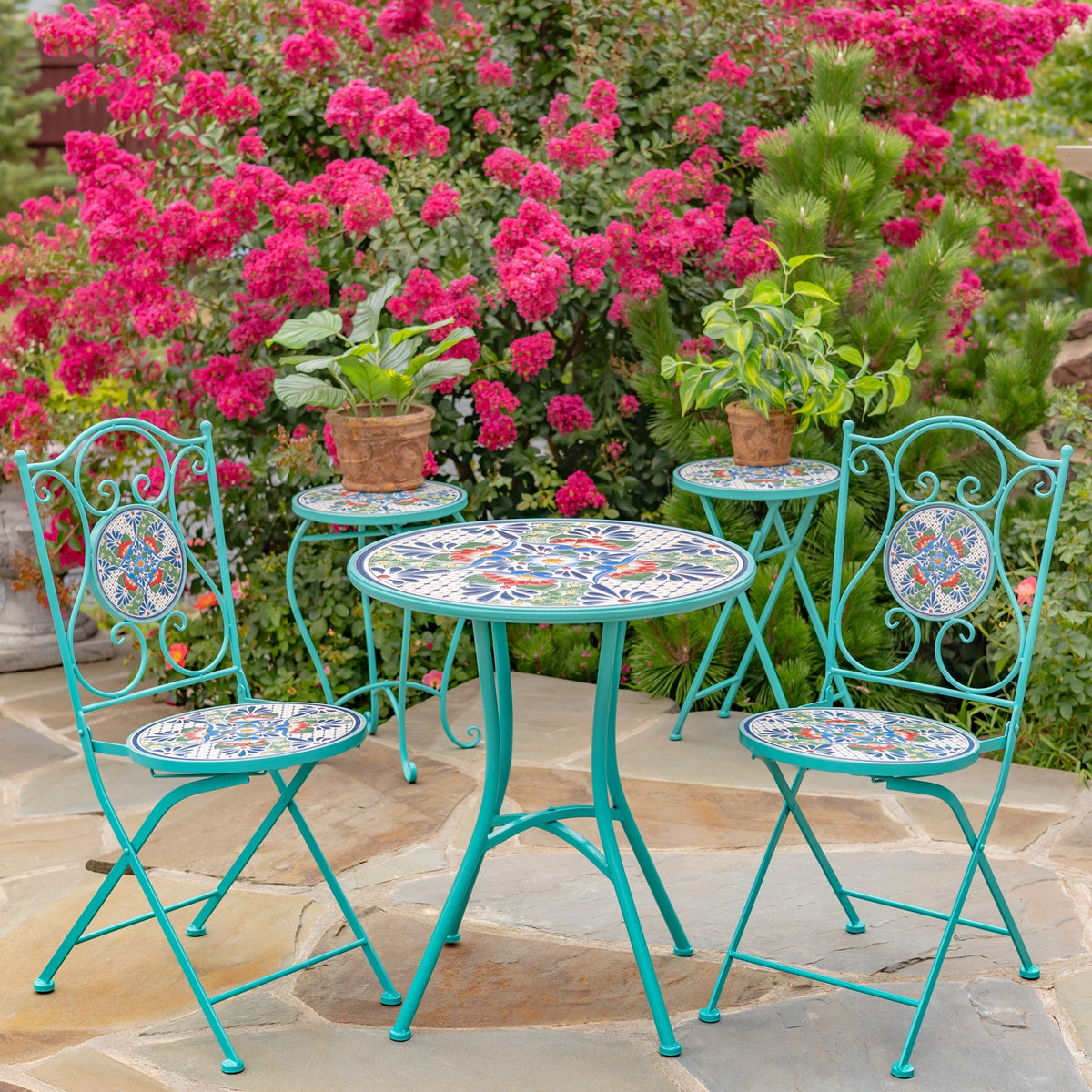 Mosaic-topped turquoise table and folding chairs arranged on a stone patio among flowering shrubs.