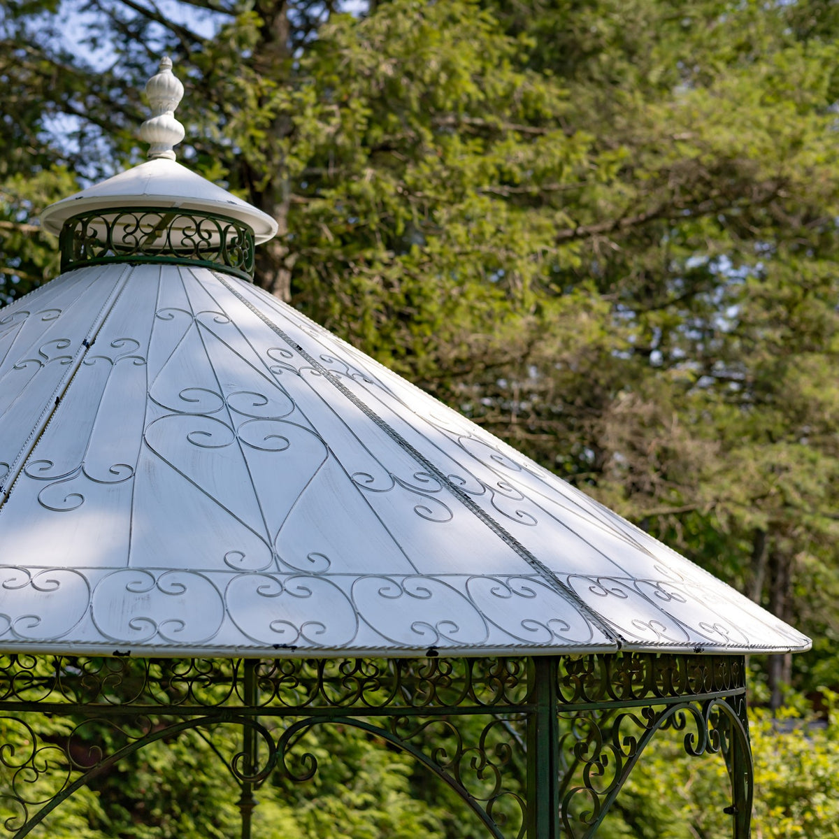 White conical gazebo roof with a small finial and decorative ironwork, set against trees.
