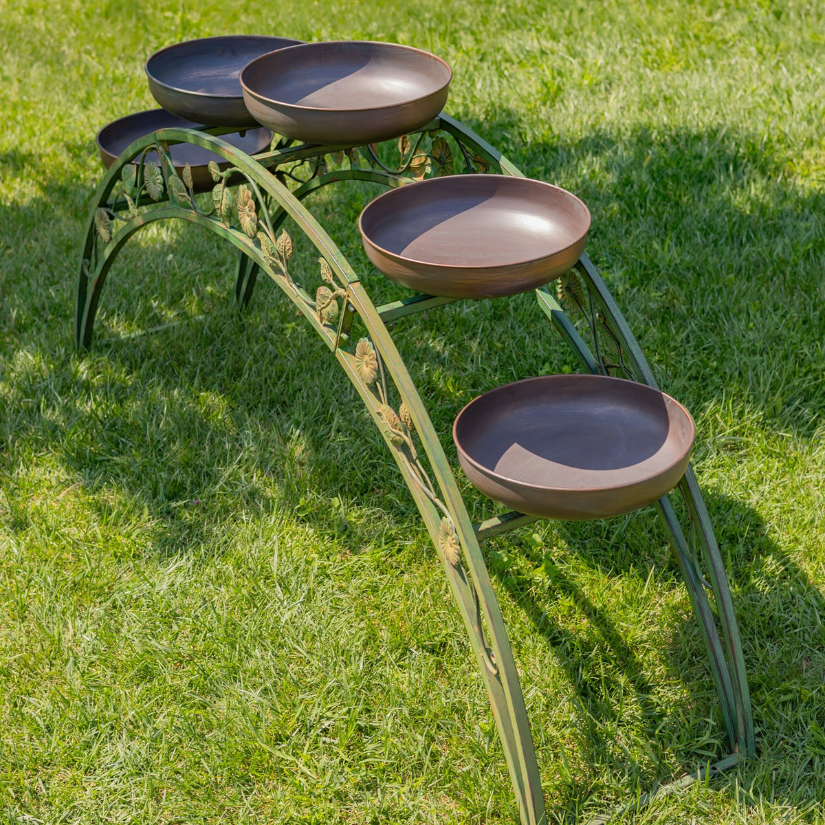 Arched green iron plant stand with four visible shallow brown planter bowls displayed on grass.