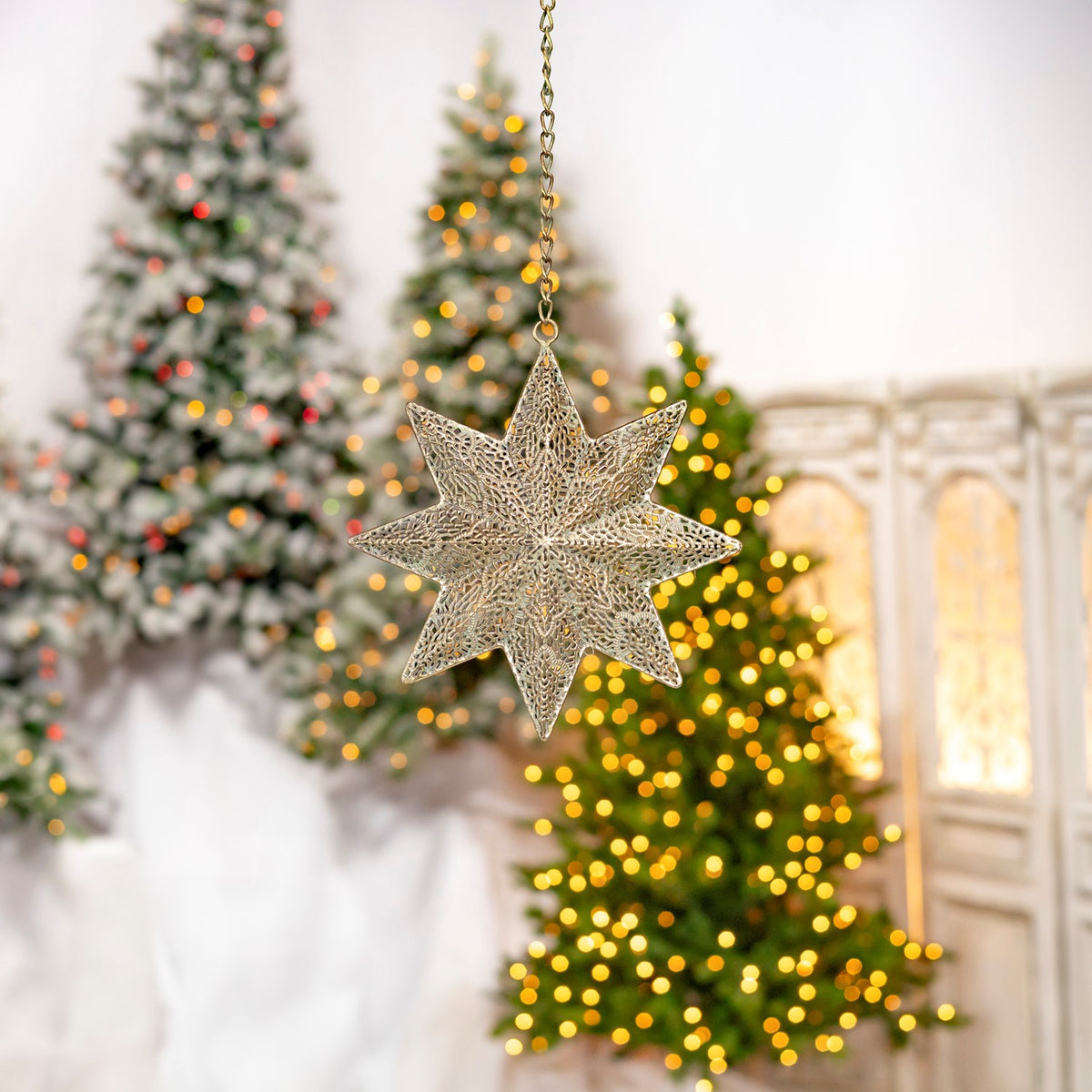 A smaller-looking openwork star ornament suspended in front of decorated trees and a lit cabinet.