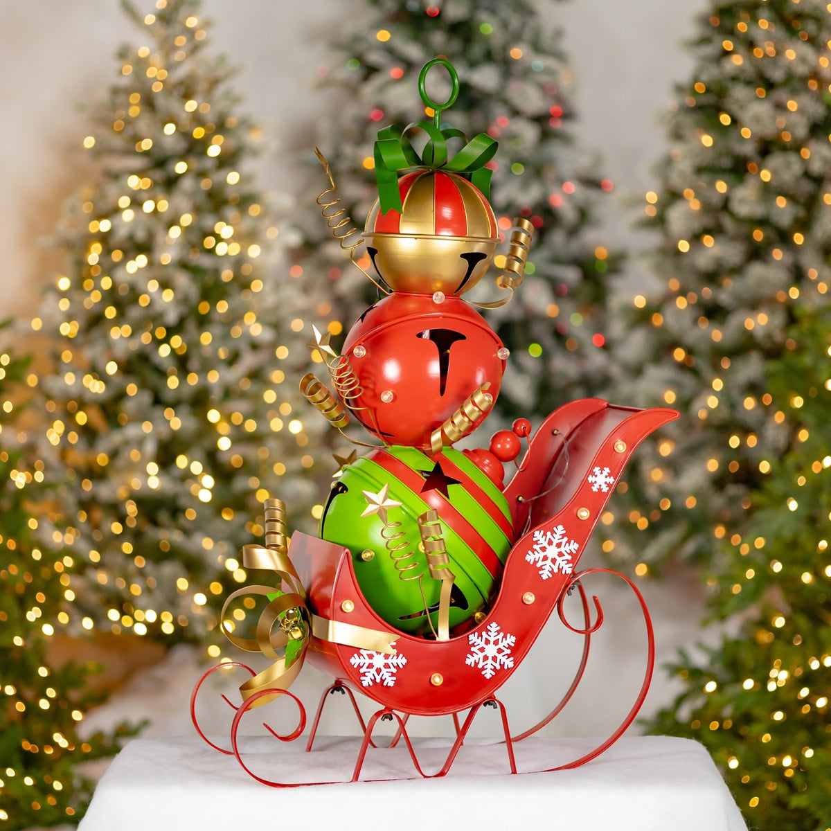 Red iron sleigh decorated with snowflakes carries colorful jingle bells, displayed on a white surface by lit trees.