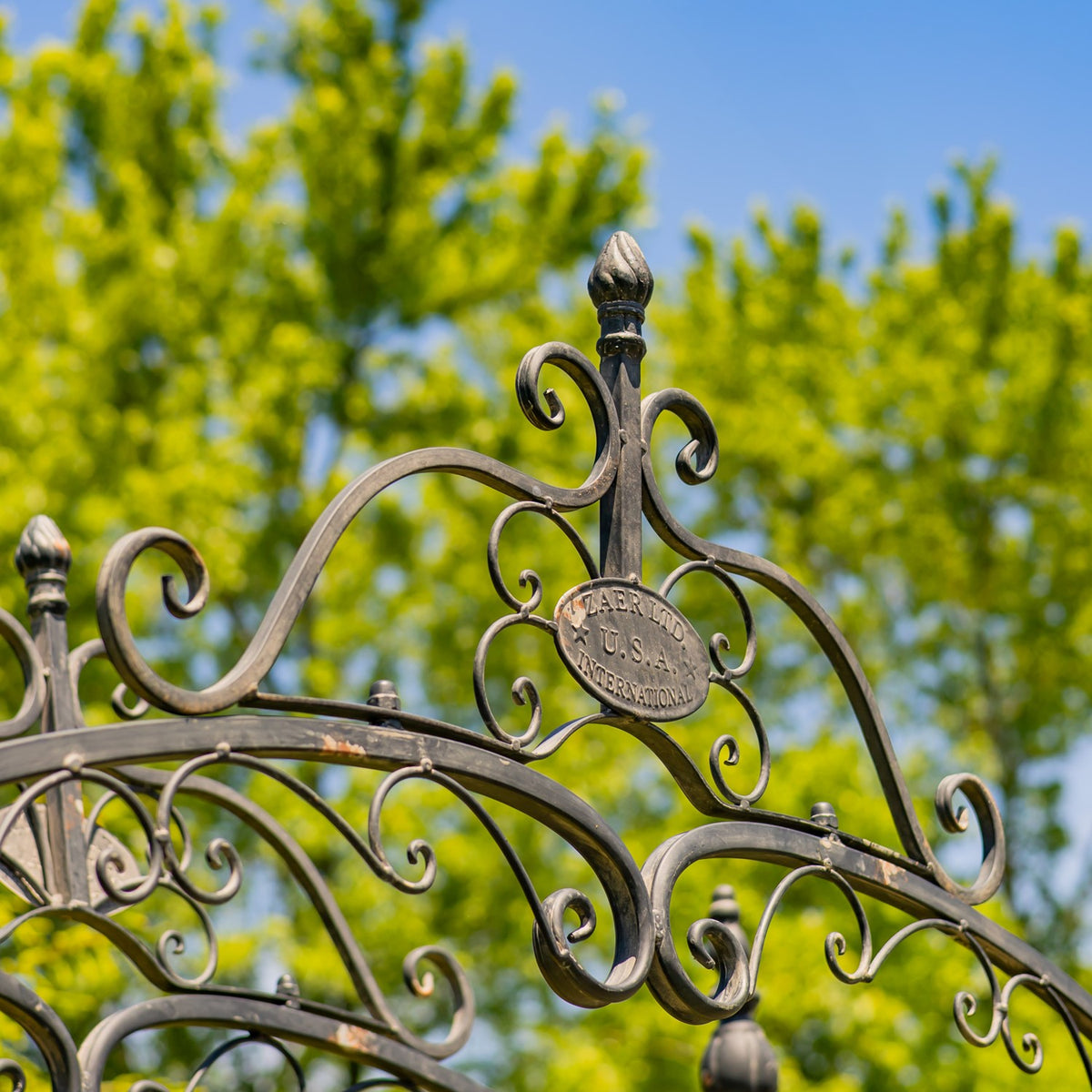 Curled iron scrolls and a small oval plaque sit beneath a pointed finial against a blue sky.
