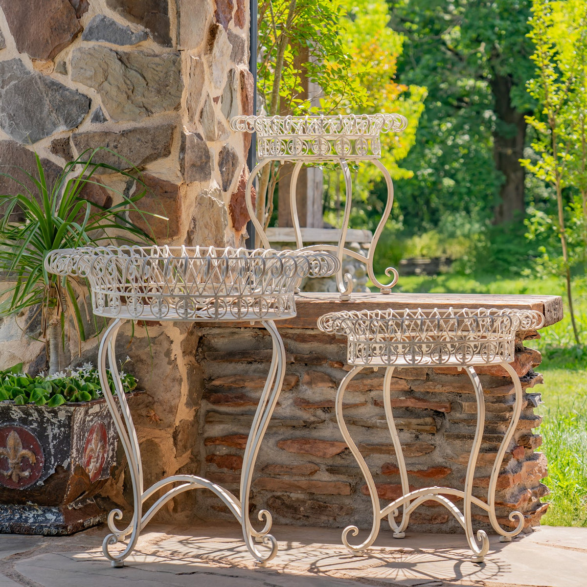 Three pale wire plant stands in different heights beside a stone wall and garden greenery.