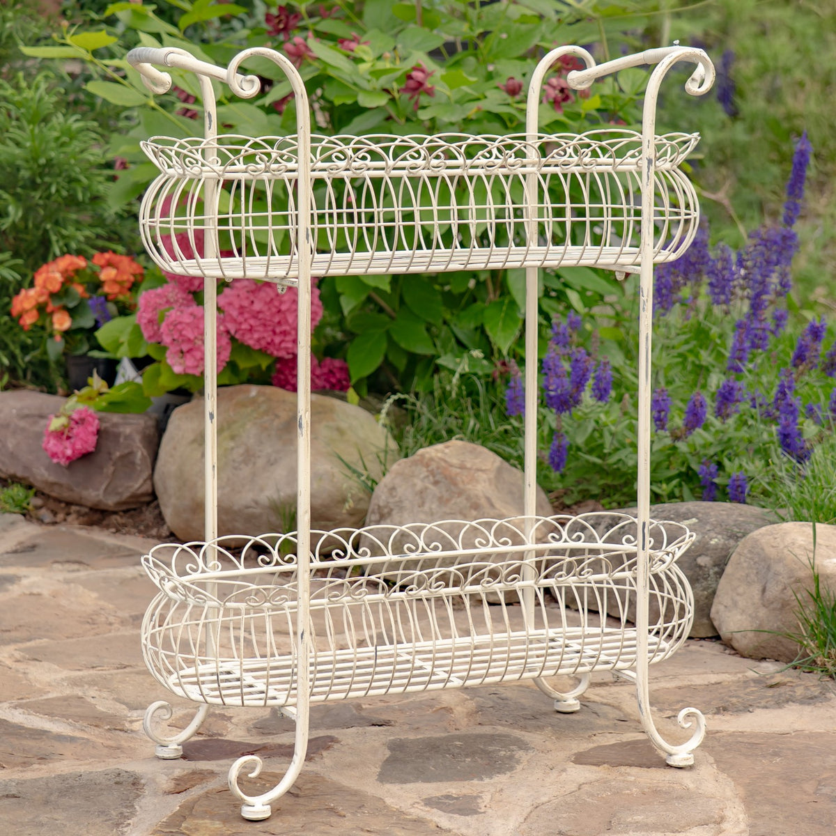 Two-tier oval wire basket plant stand with curled handles and four curved feet on a garden path.