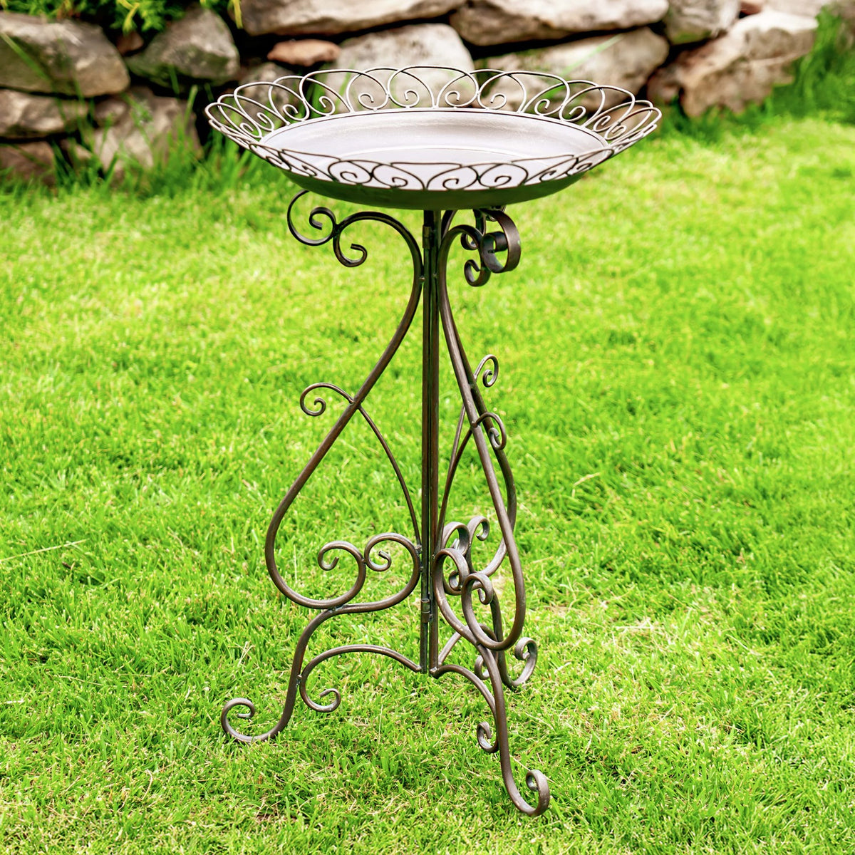 Round birdbath with a decorative scrollwork rim and slender folding stand, shown on a grassy lawn beside a stone wall.