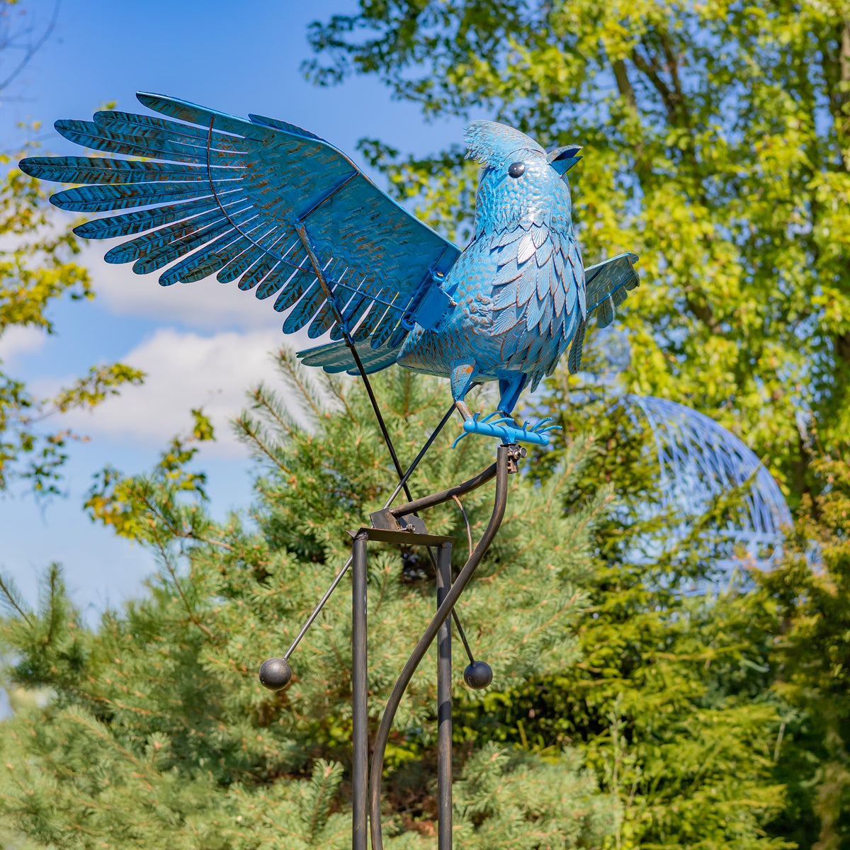Blue jay sculpture with wings spread perches on a tall iron stake with hanging counterweights in a garden.