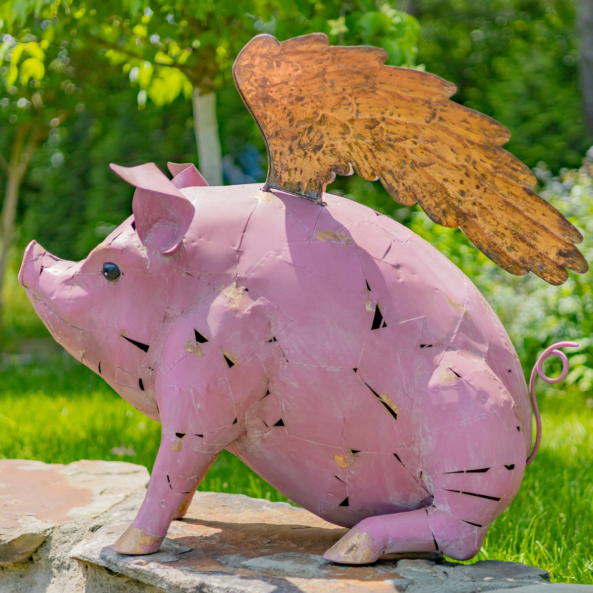 Side view of a pink metal pig statue with a large golden wing and curled tail in a garden.