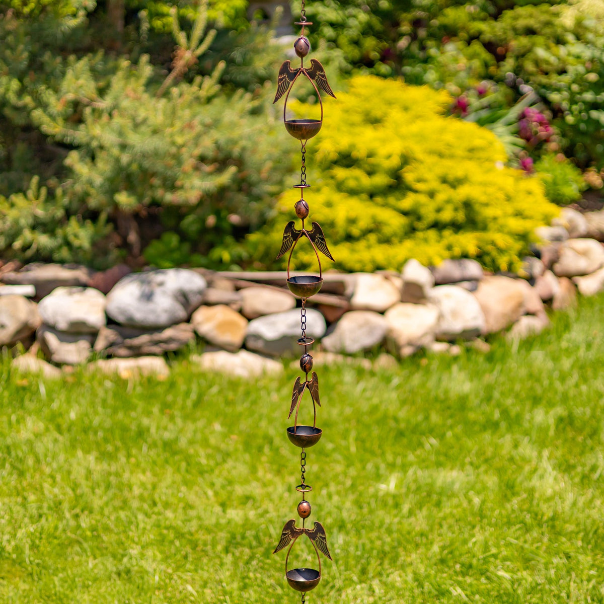 A long rain chain with repeating angel figures and bowl-shaped cups hangs in a garden setting.
