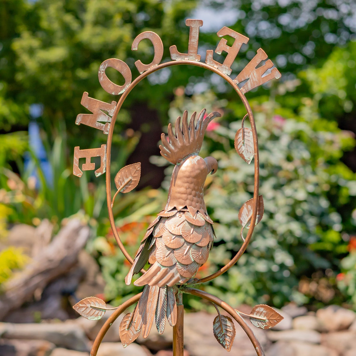 Metal cockatoo garden stake with raised wings inside a circular “WELCOME” frame, surrounded by greenery.