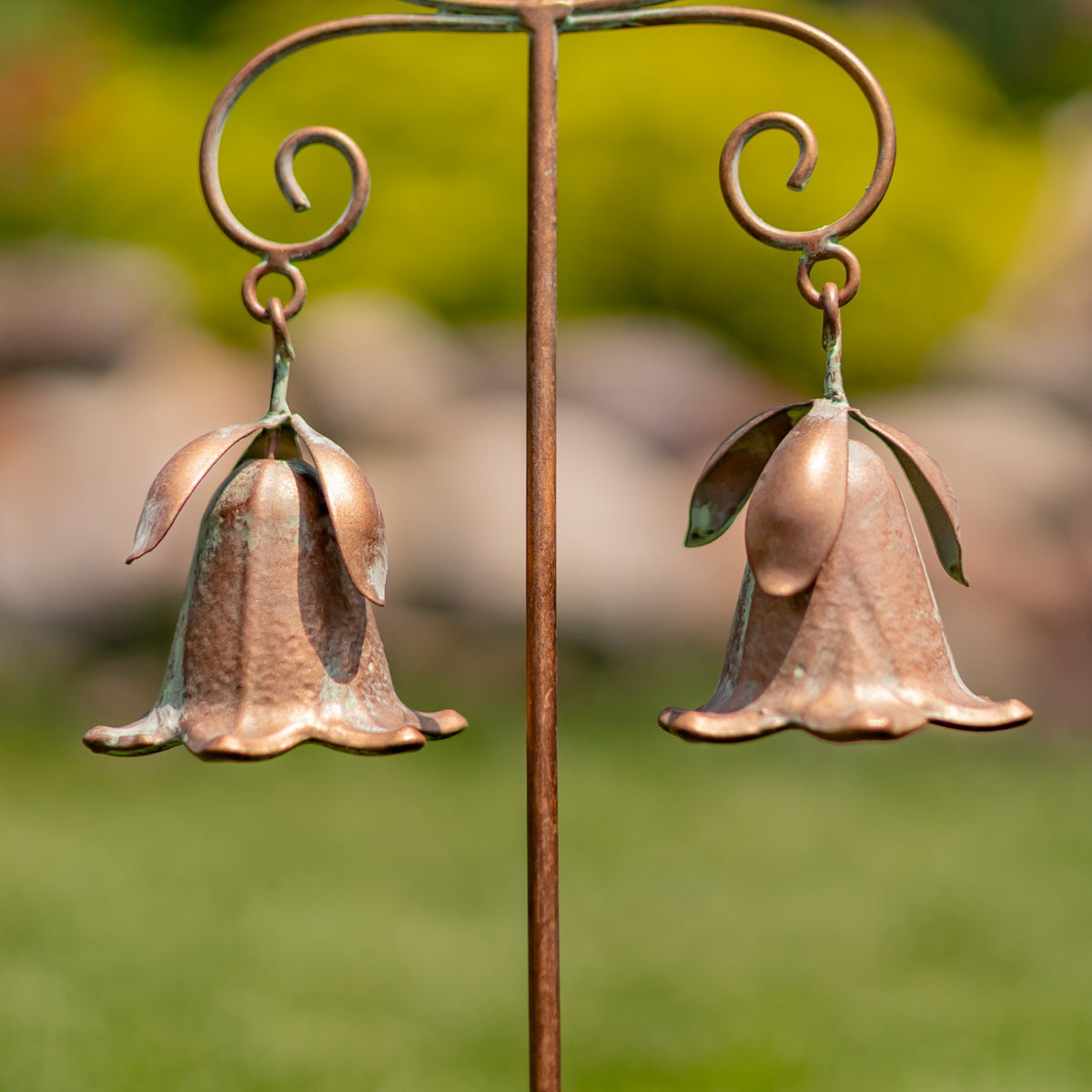 Two copper-toned flower-shaped bells hang from curled hooks on a garden stake against a grassy background.