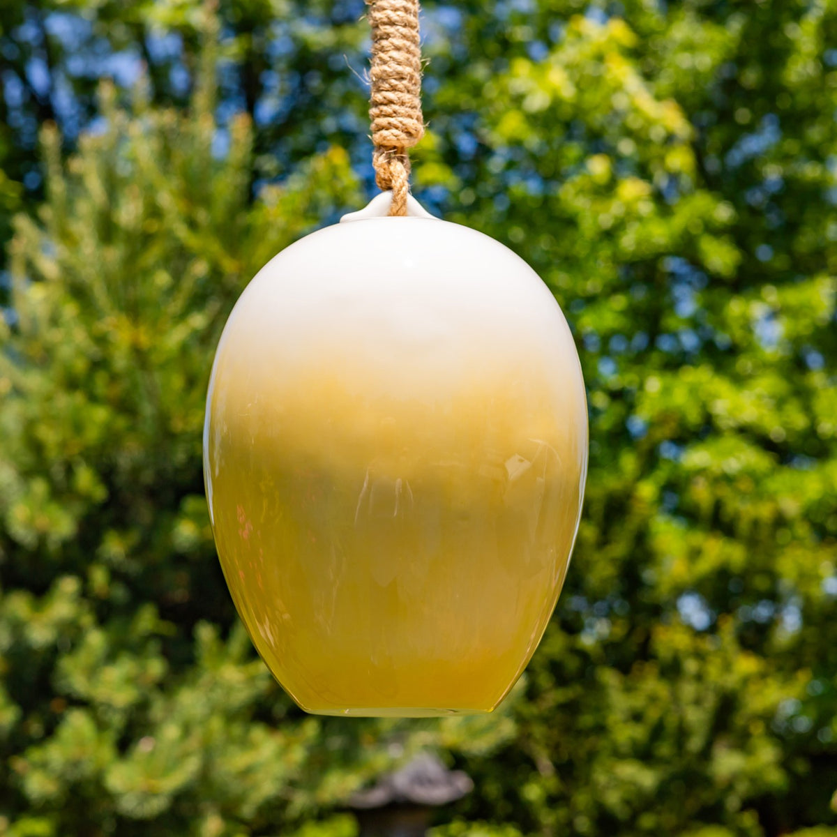 Plain yellow-to-white back of the egg-shaped birdhouse hanging outdoors by a rope.