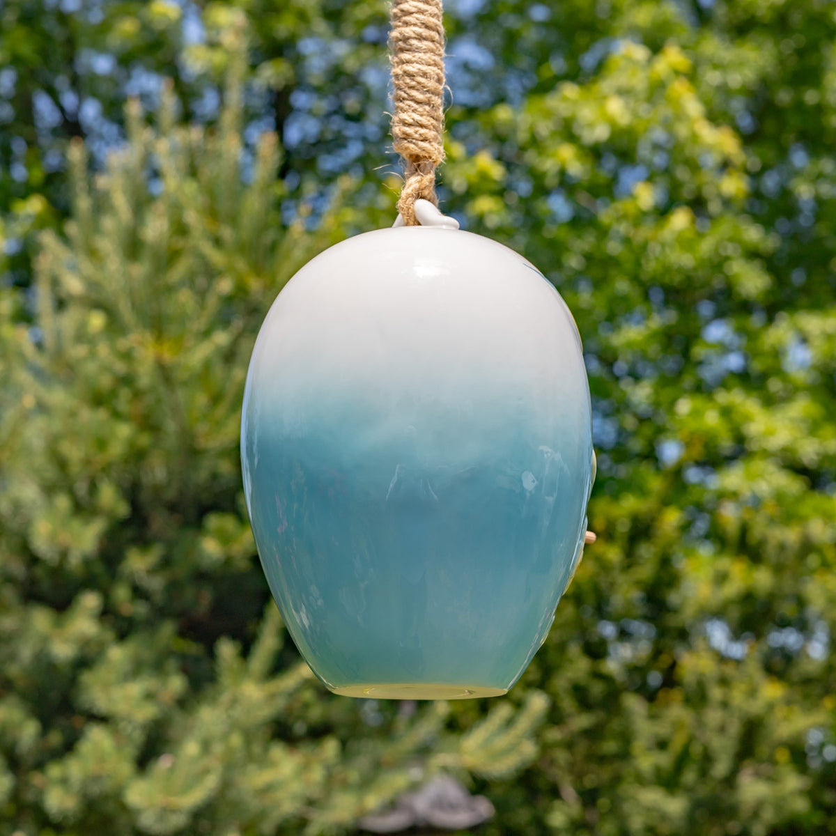 Plain white-to-blue egg-shaped birdhouse hanging by a thick rope against a leafy garden.