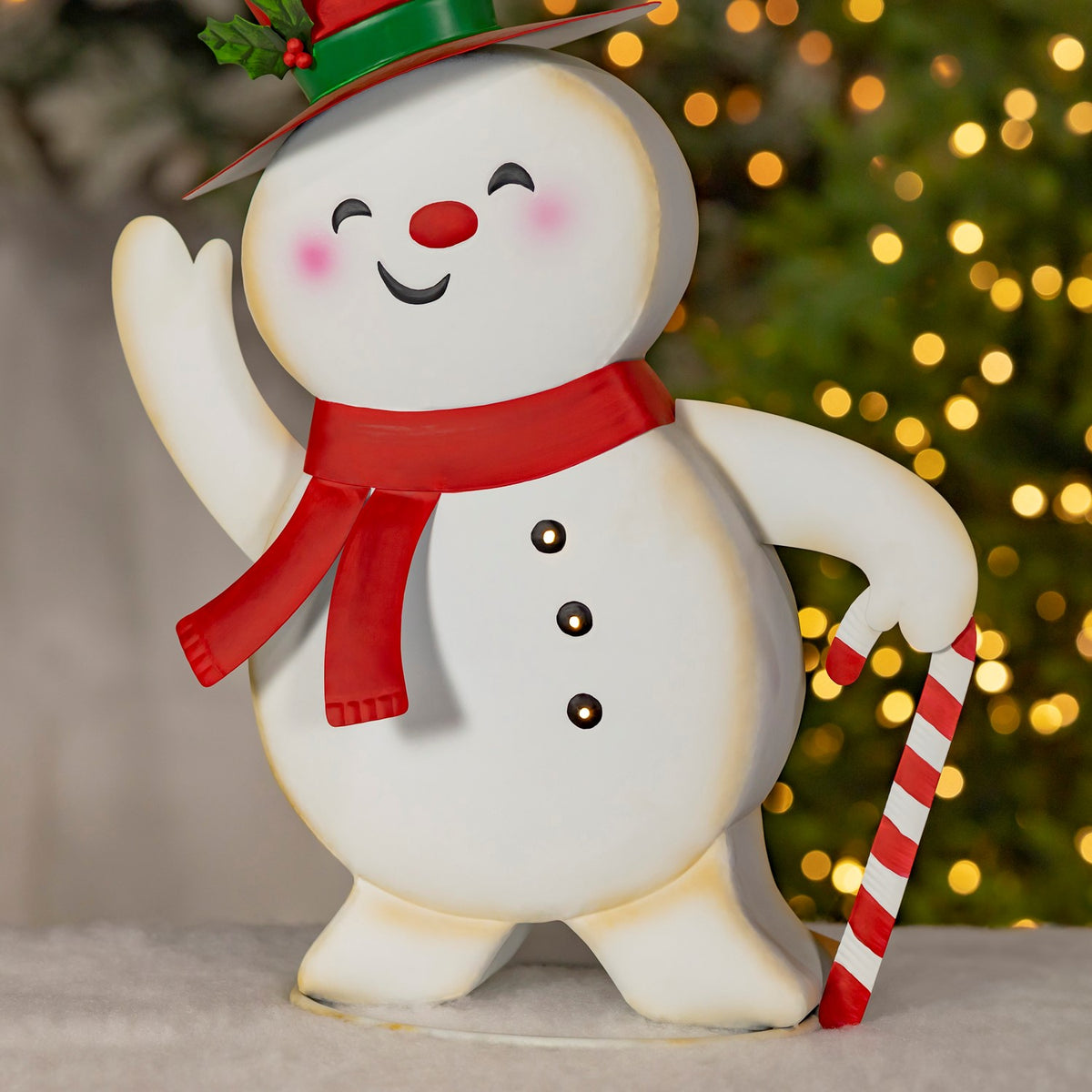 Full snowman figurine waving and holding a red-and-white candy cane beside a Christmas tree.