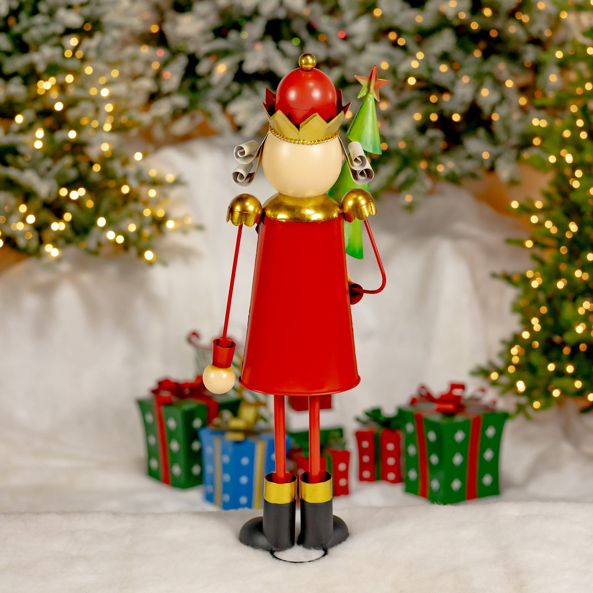 Red-coated nutcracker figurine with a red hat, gold crown, dangling ornaments, and a small tree against a festive backdrop.