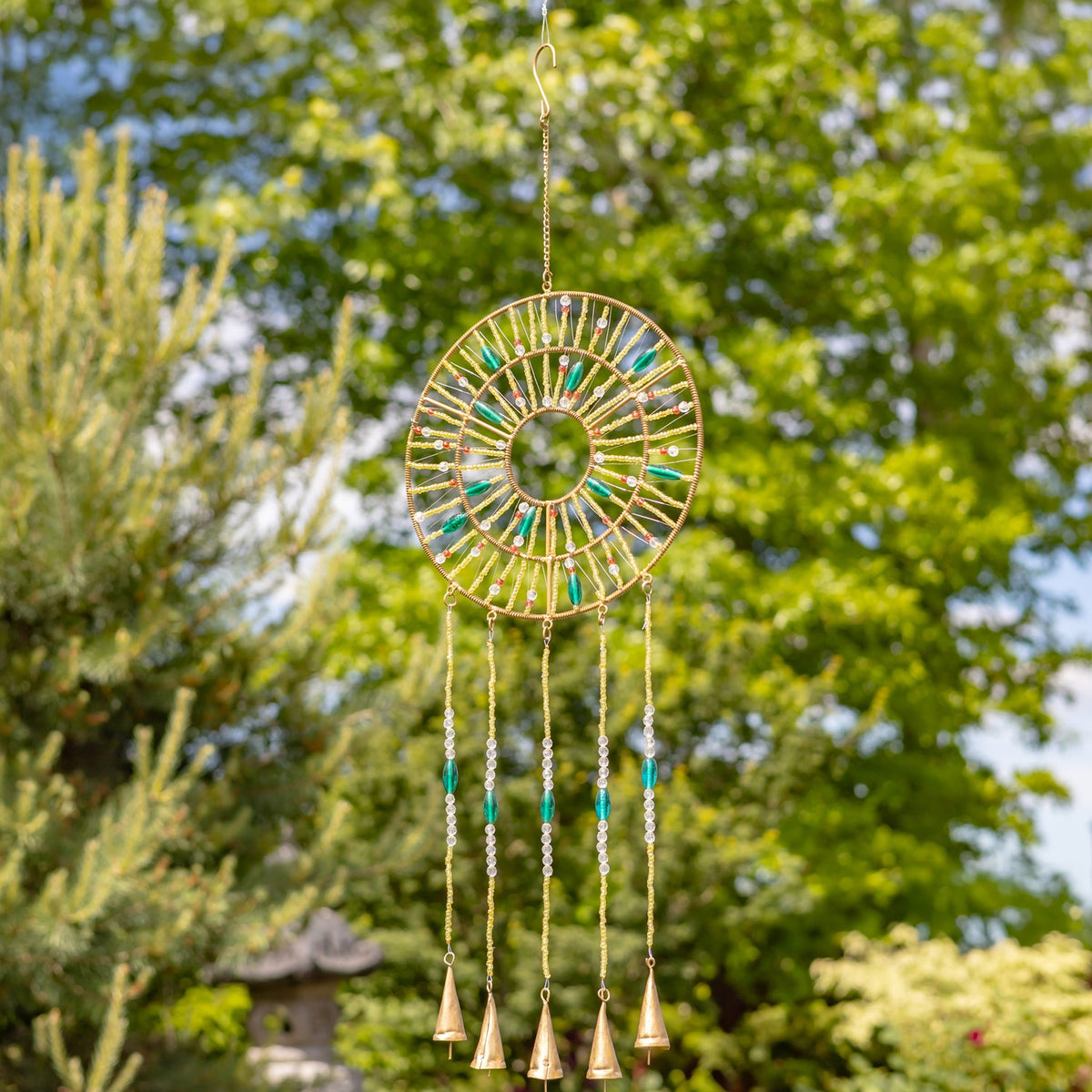 Round beaded wind chime with turquoise accents and five hanging bells among green trees.
