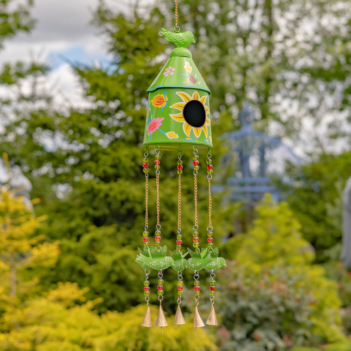 Light green birdhouse wind chime decorated with flowers, hanging against leafy garden trees.