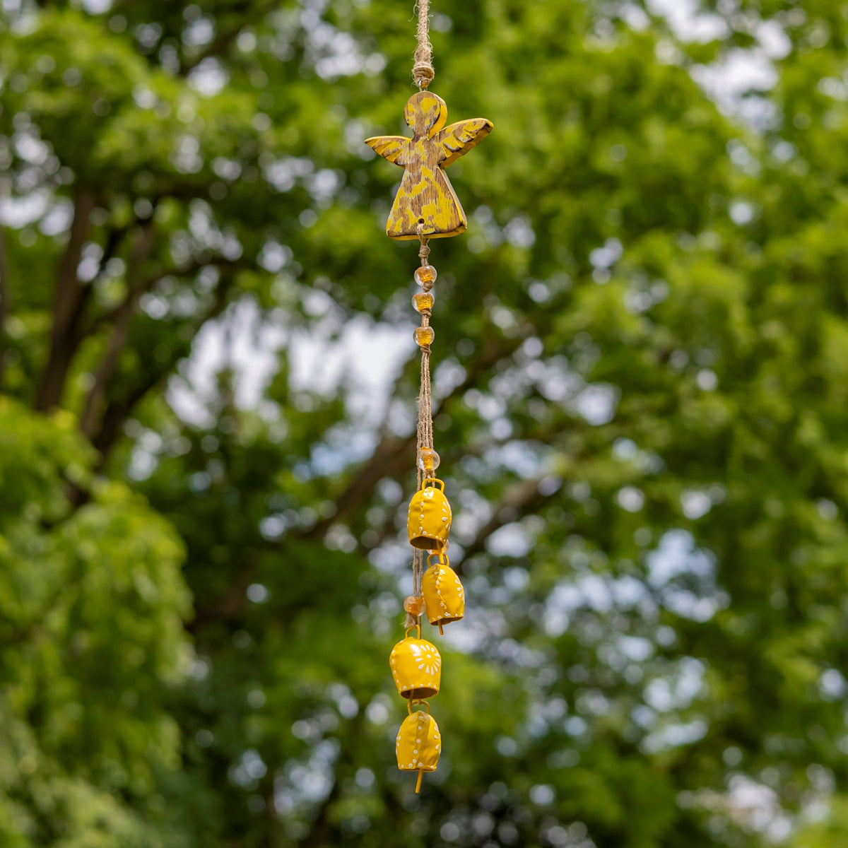 Yellow angel wind chime with dangling bells hanging against a leafy green background.