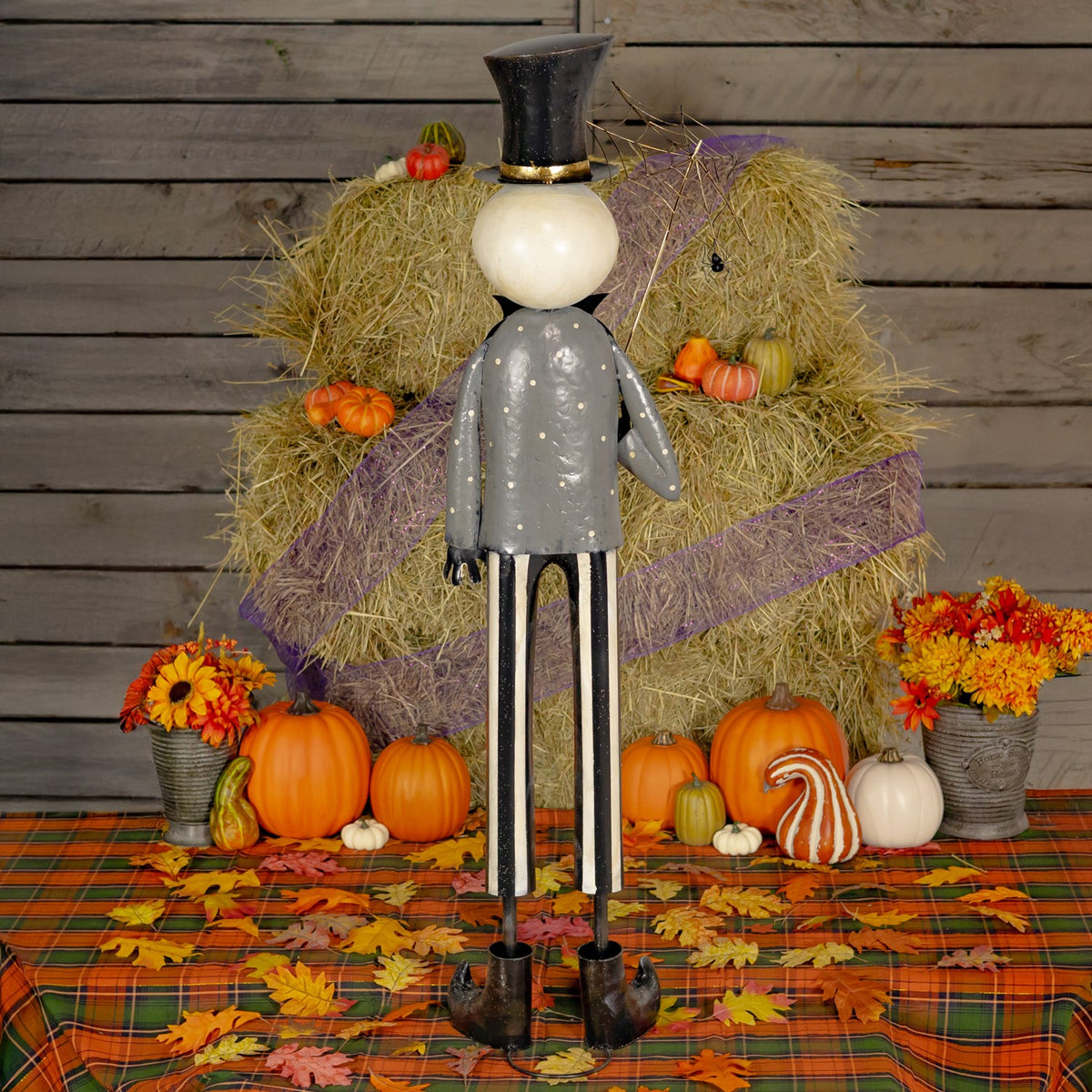Full-length skeleton figure in a tall black hat and gray coat, posed before hay bales, pumpkins, and autumn leaves.