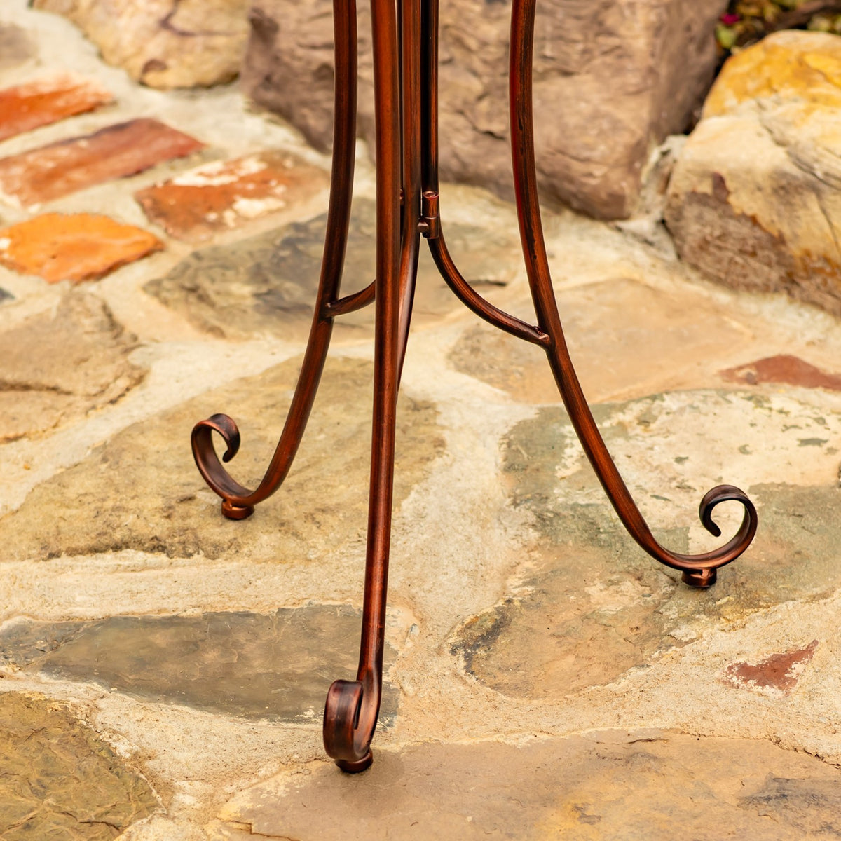 Curved metal legs of the birdbath stand end in scroll-shaped feet on a stone patio.