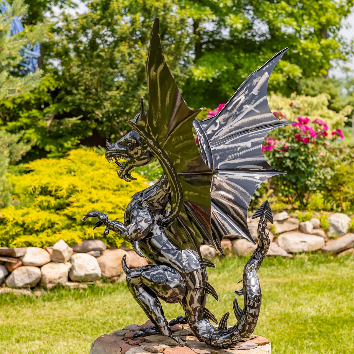 Full view of a shiny metal dragon standing upright on a stone base with wings spread in a garden.