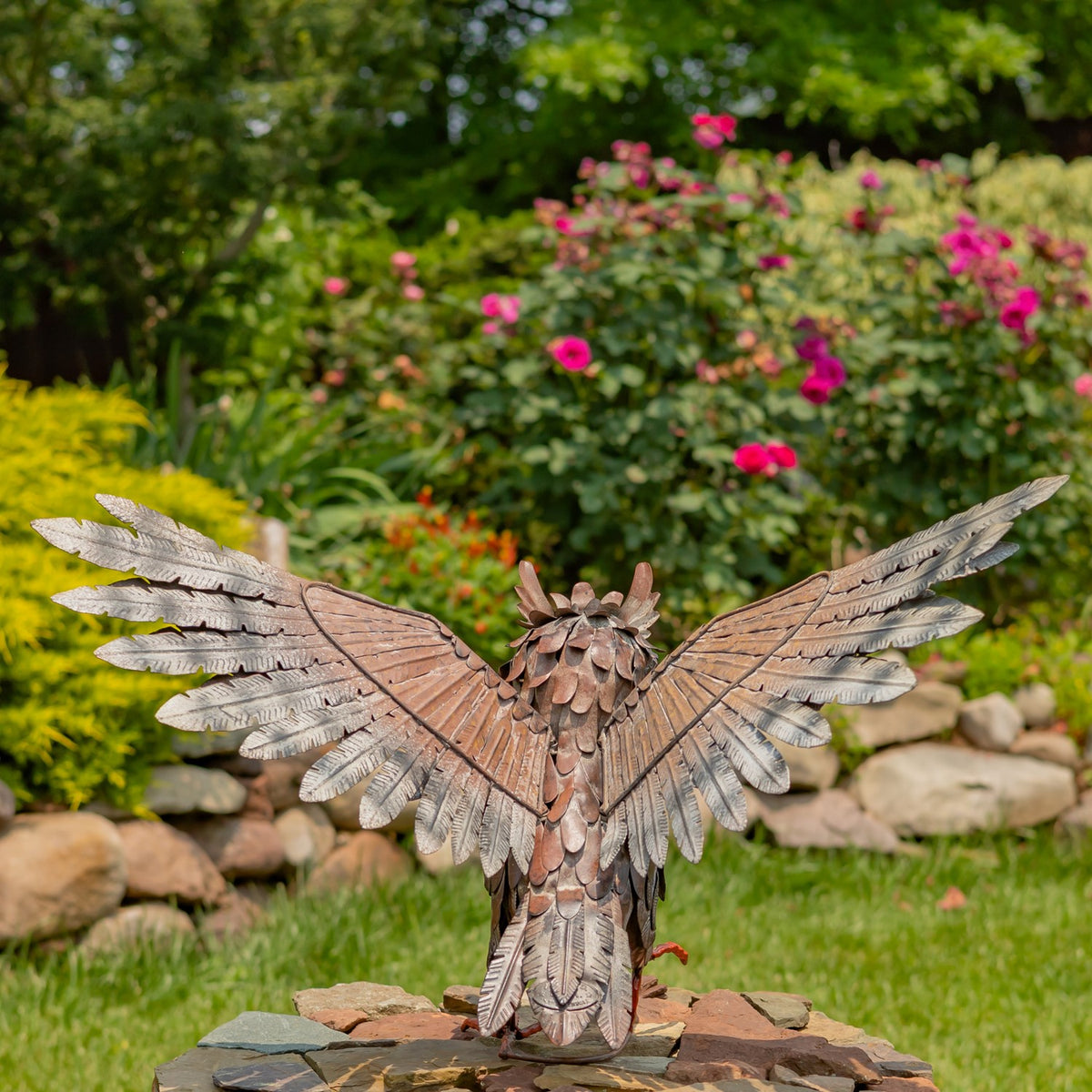 Metal owl seen from behind with broad wings spread, standing in a garden among flowers and stones.