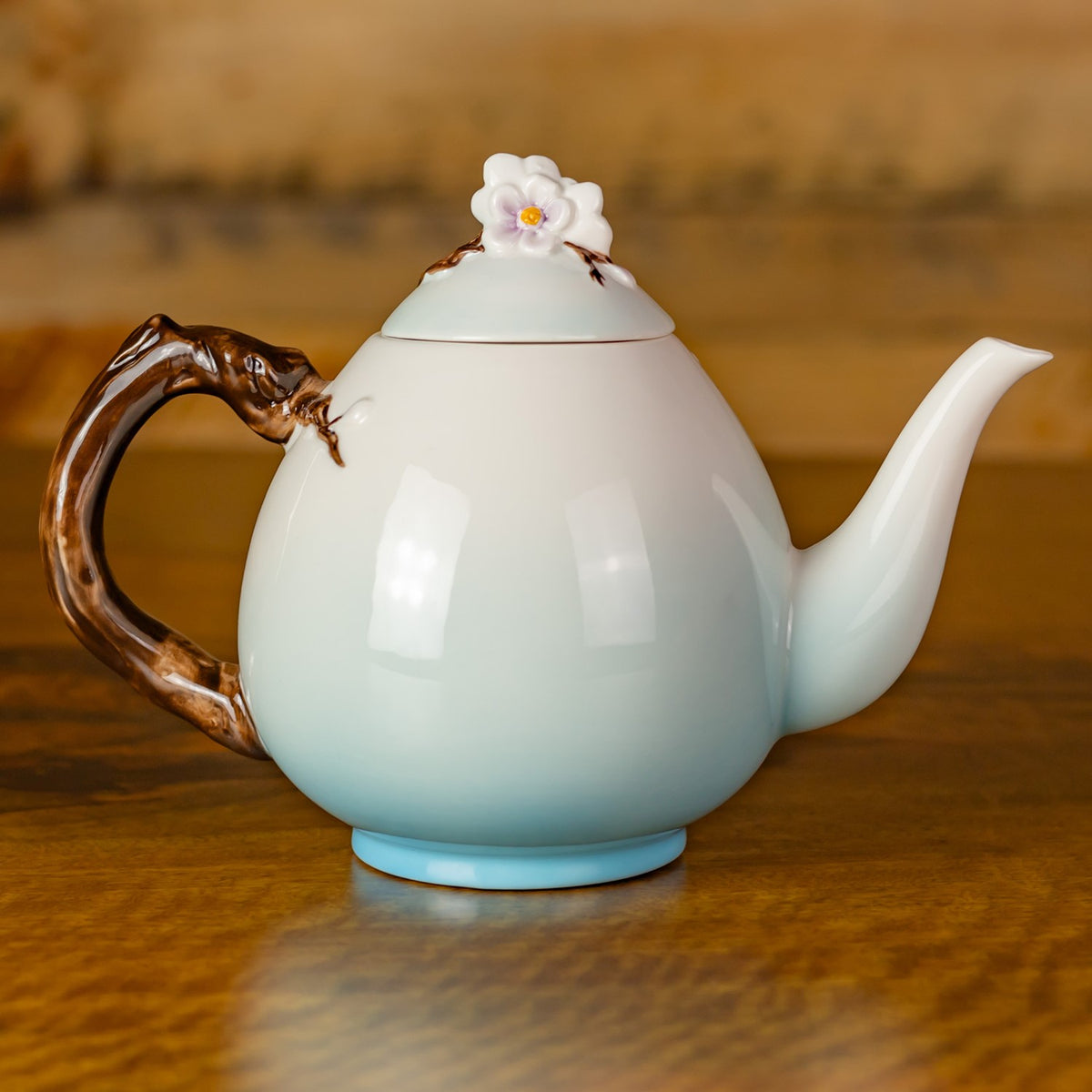 Plain pale blue-white back of the teapot, showing its brown branch-like handle, curved spout, and flower-topped lid.