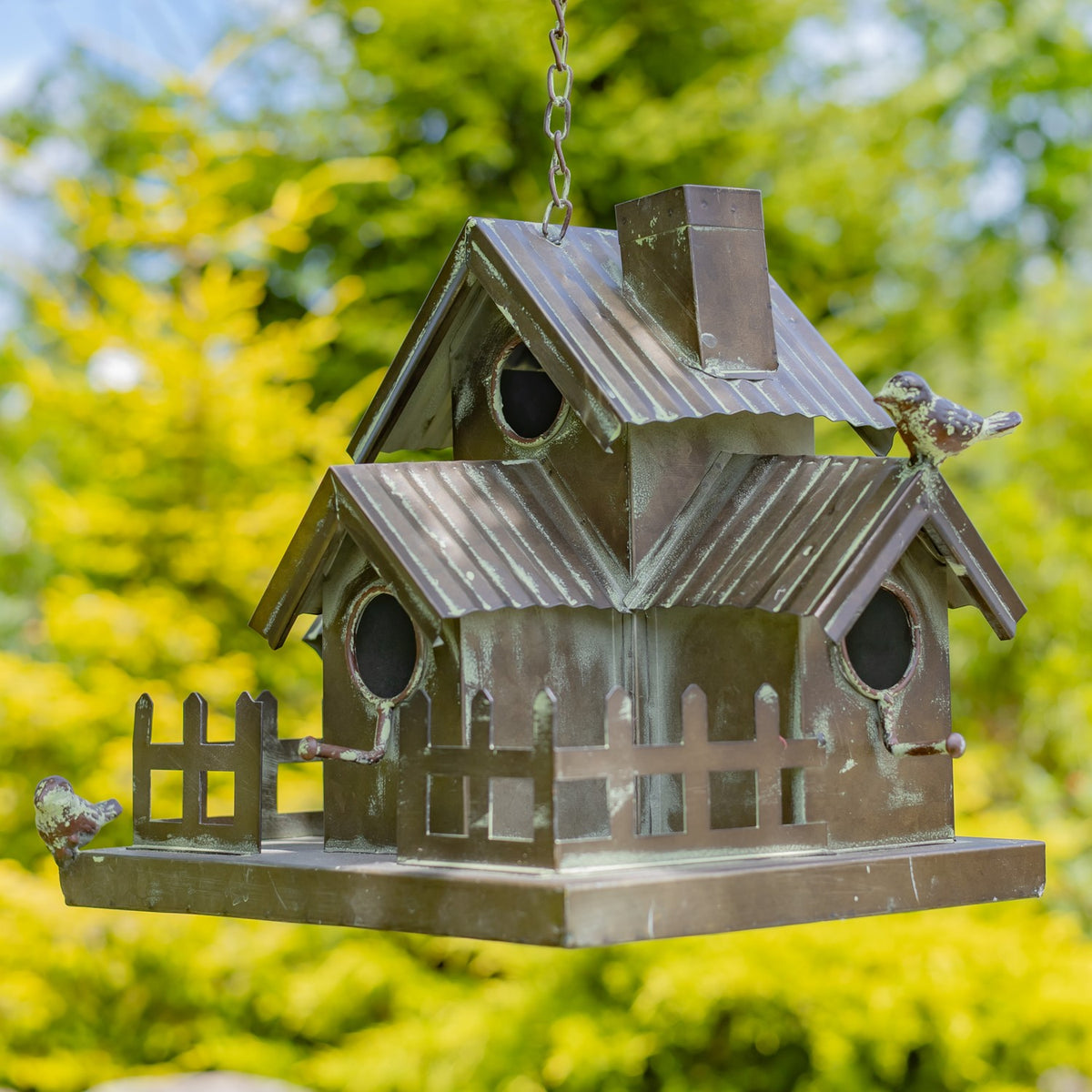 A multi-section birdhouse hangs from a chain, showing several entry holes, a chimney, rooflines, and fence details.