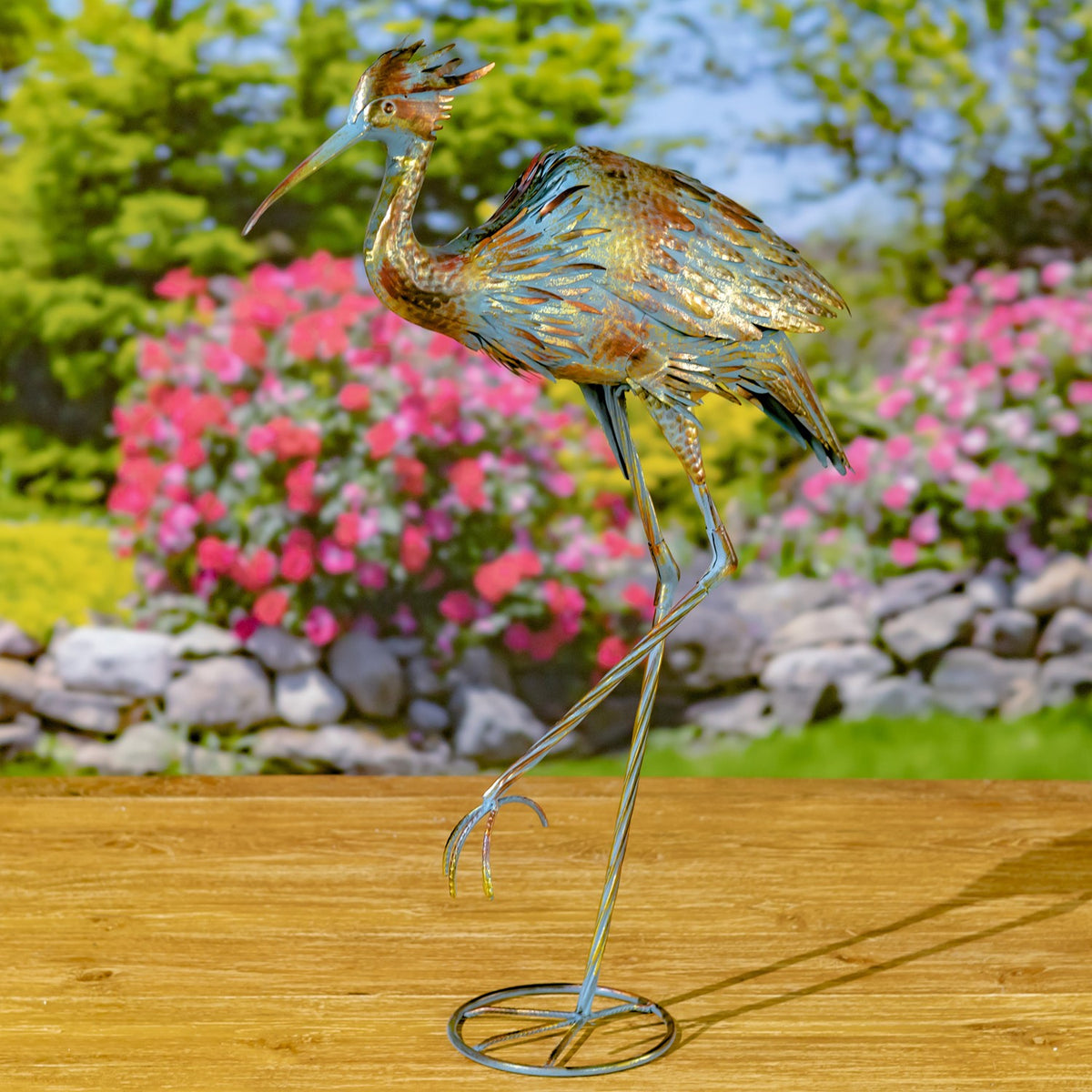 Side view of a metal heron figurine on a circular wire base, posed against a garden backdrop.