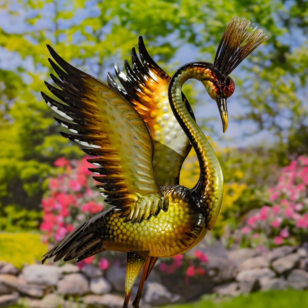 Metallic heron figurine with one wing raised, its head bent toward the ground in a garden setting.