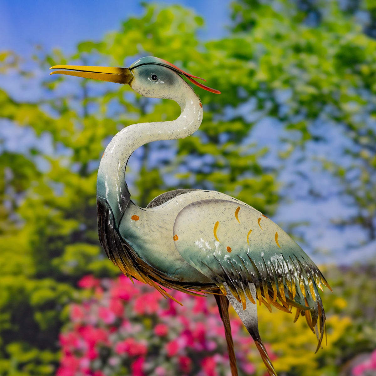 A metal crane figurine seen from the other side, its long beak pointing left against a leafy garden backdrop.