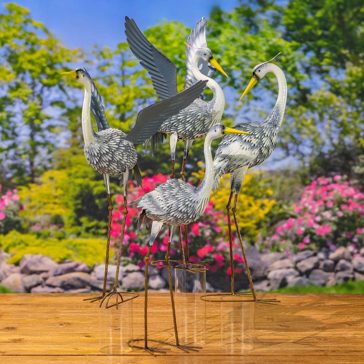 Four metal heron figurines in varied poses stand together on a wooden surface against a flowering garden.