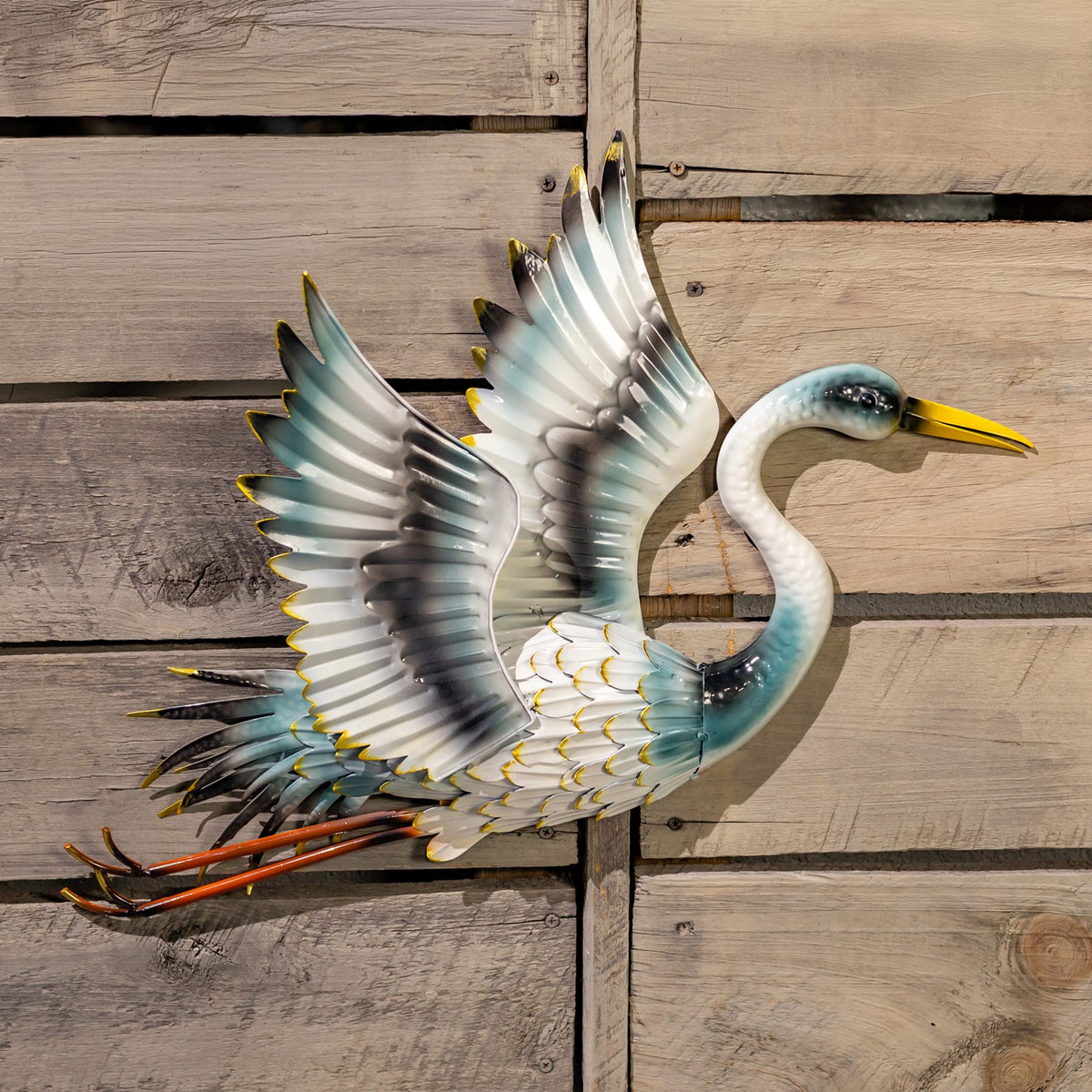 A metal crane ornament faces right with spread wings, a curved blue neck, yellow beak, and trailing legs.