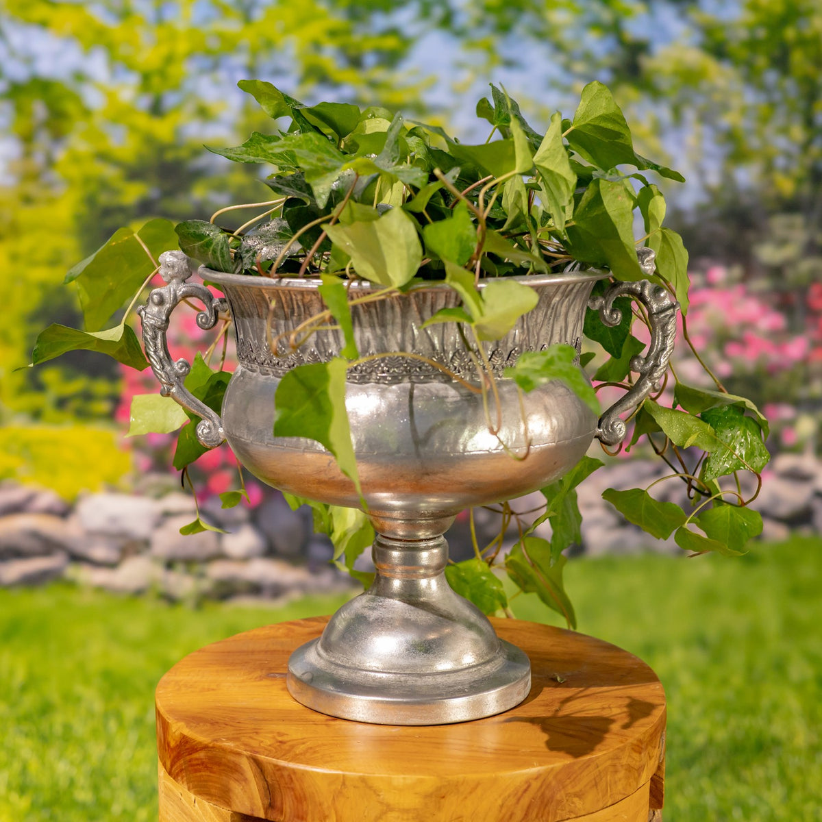 Frosted silver urn with ornate handles holding leafy greenery on a wooden stand.