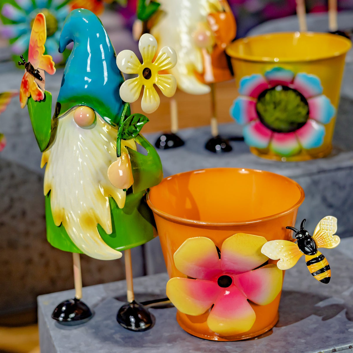A blue-hatted gnome stands next to an orange pot with a pink flower and a small bee decoration.