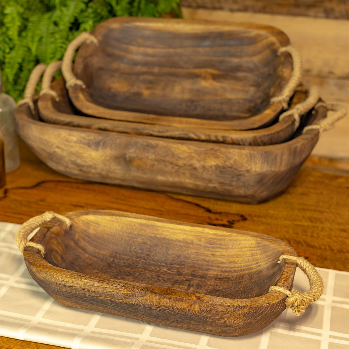 Several oval wooden trays with rope handles, nested on a table beside a matching tray in front.