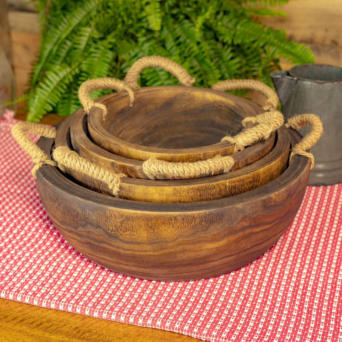 Four round wooden trays with rope handles are nested together on a red checked cloth.