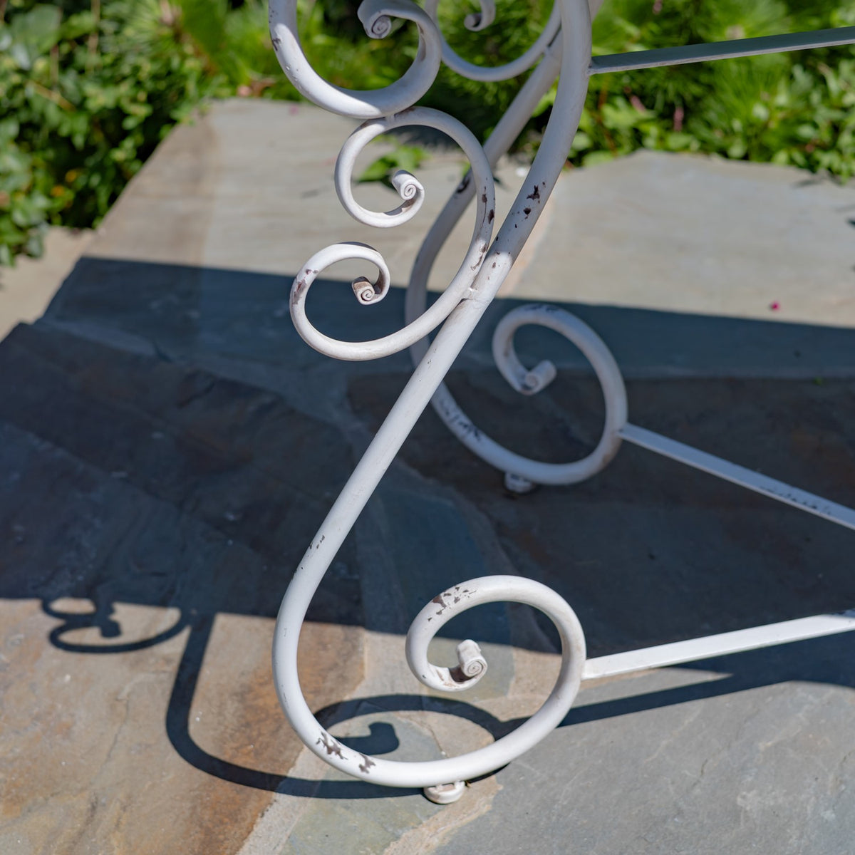 White iron table legs with curled scrollwork and scattered paint chips against garden greenery.