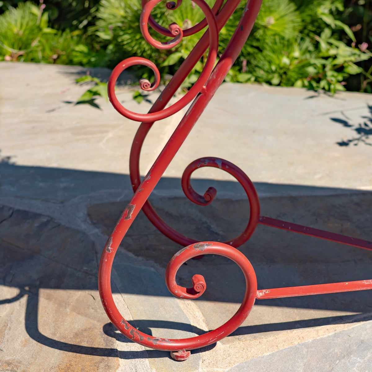 Close-up of a red iron table leg with curled scrollwork and patches of worn paint against a stone patio.