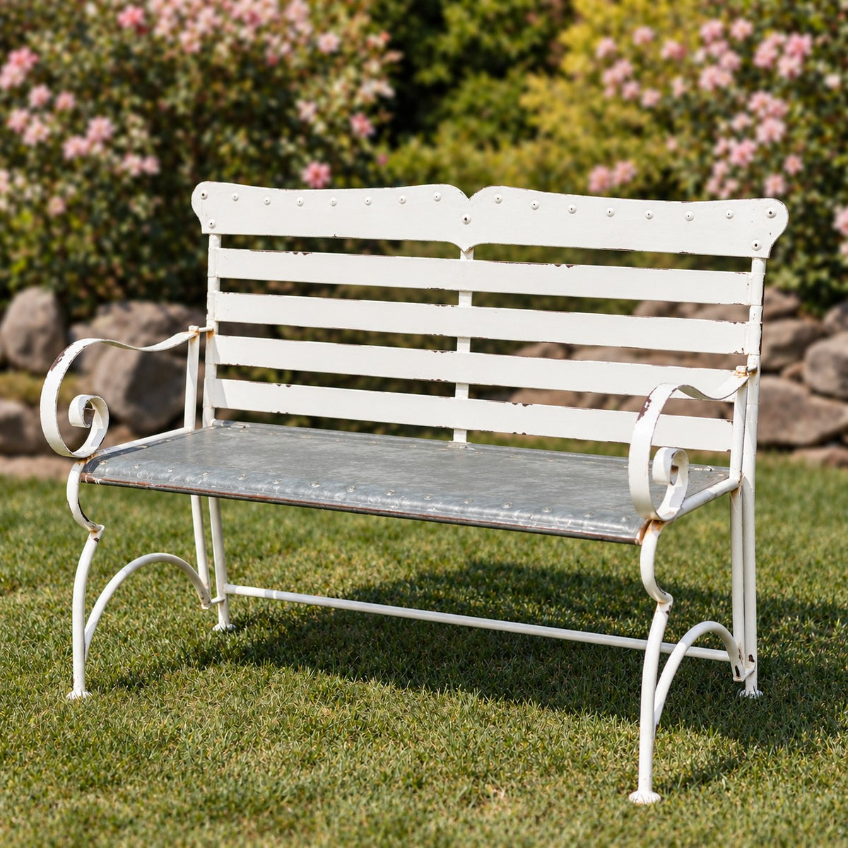White metal garden bench with a slatted back and curled arms on a lawn.