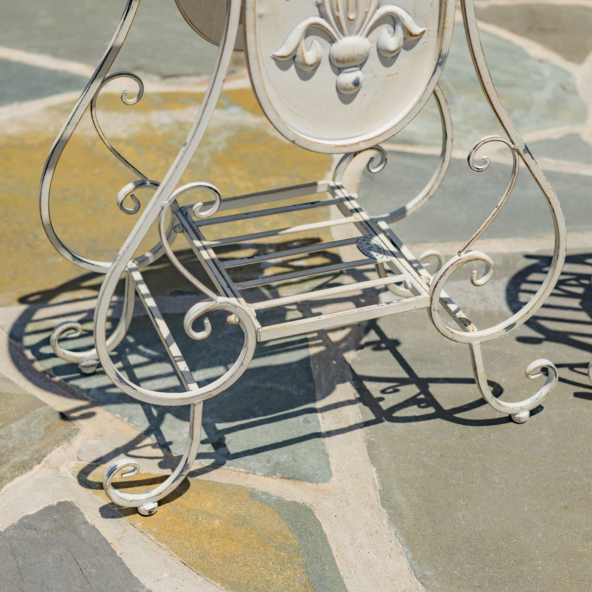 Curving ivory metal legs and a slatted lower shelf cast shadows on a stone patio.
