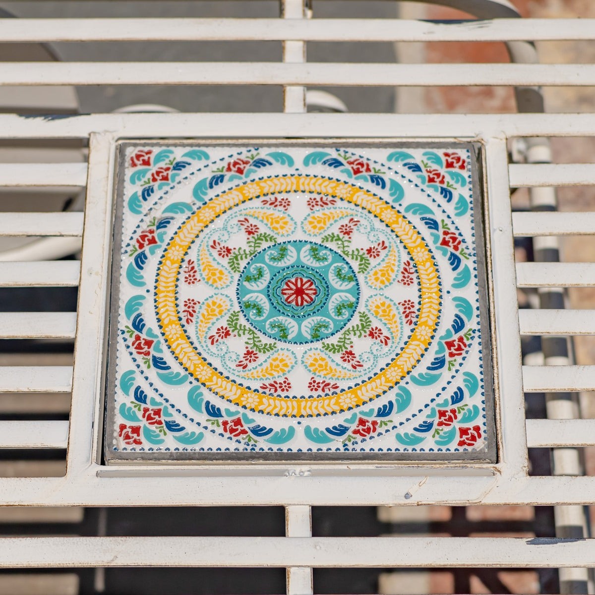 Close-up of a square tabletop mosaic with a colorful circular floral pattern set among white metal slats.