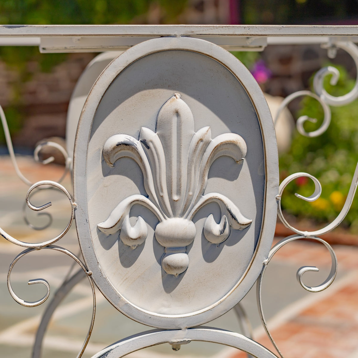 Raised fleur-de-lis ornament on an oval panel, framed by pale metal scrolls against a garden backdrop.