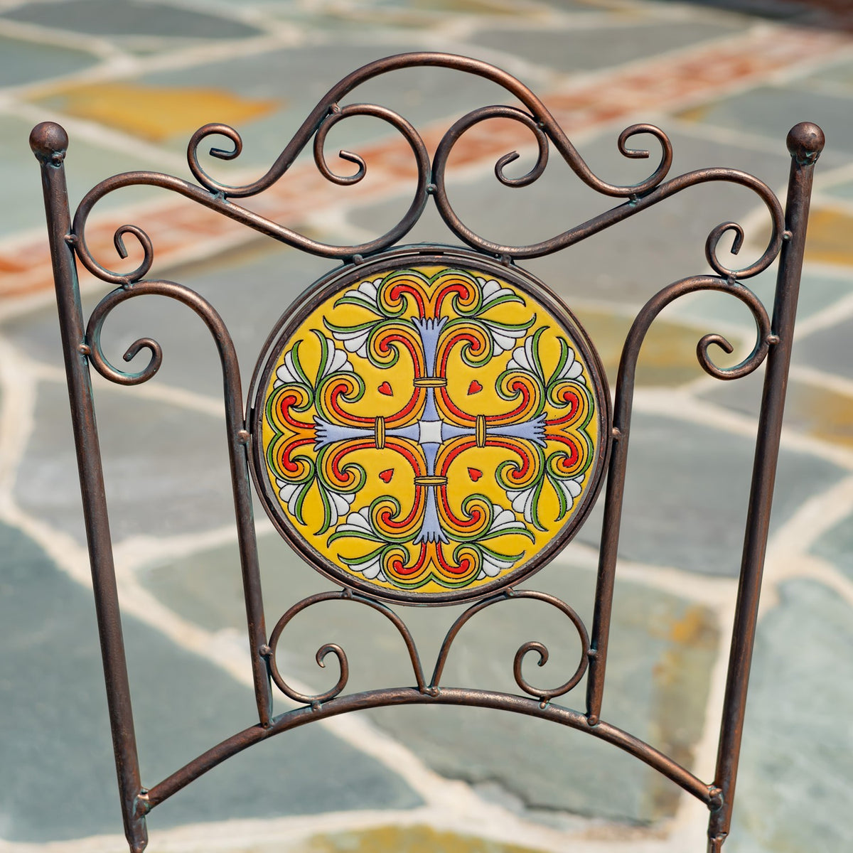 A dark metal chair back with curled scrollwork surrounding a round yellow, red, green, and blue mosaic panel.