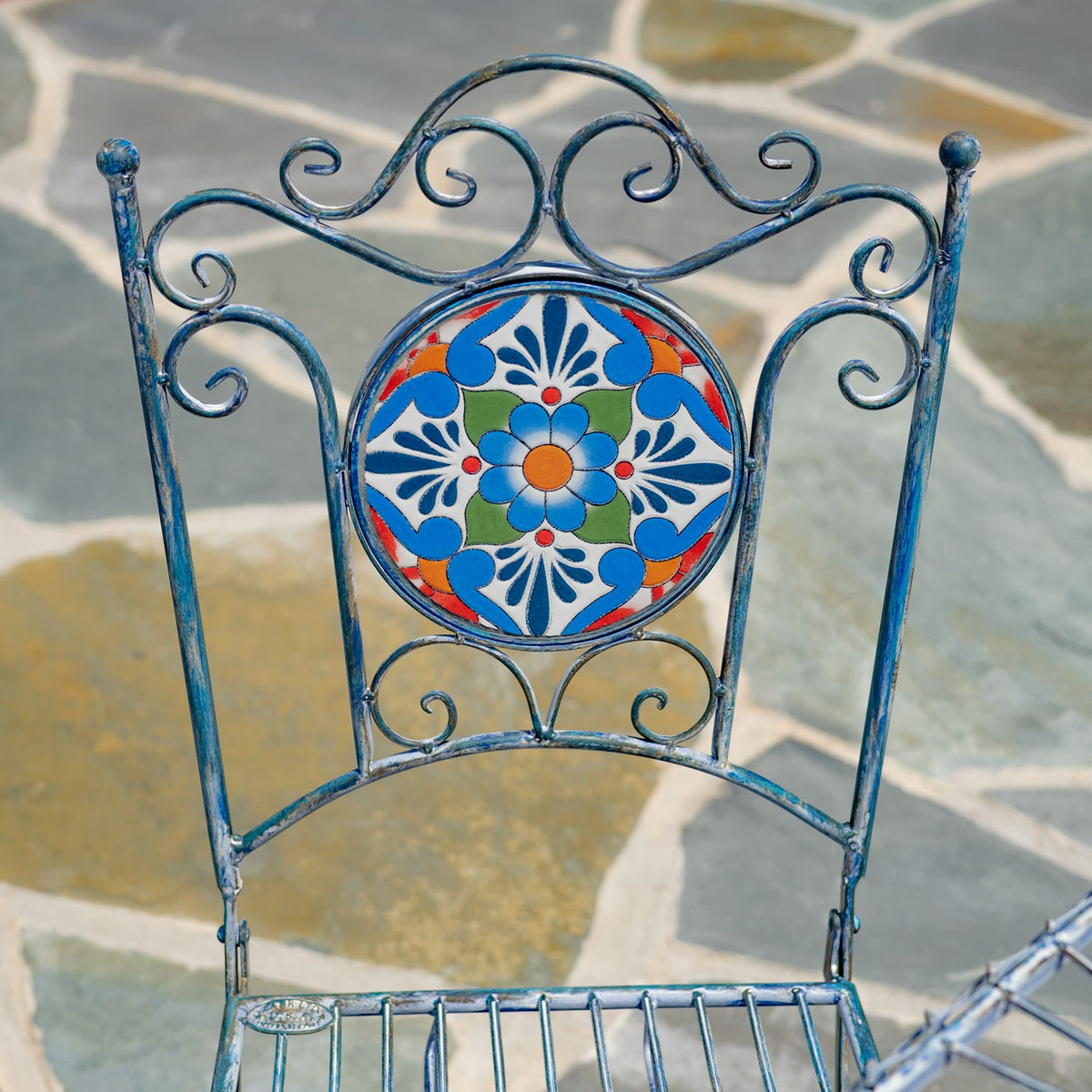 Wrought-metal chair back with curled scrollwork surrounding a round, colorful floral mosaic panel.