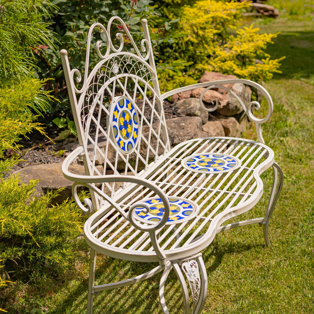 White metal garden bench with blue and yellow mosaic accents among rocks and greenery.