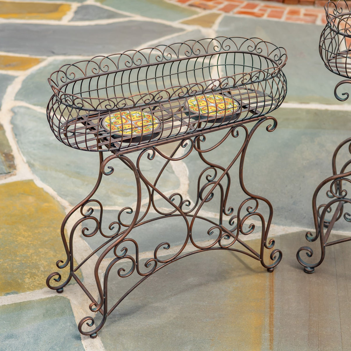 Oval metal plant stand with an openwork scroll design and two colorful mosaic panels on its top shelf.