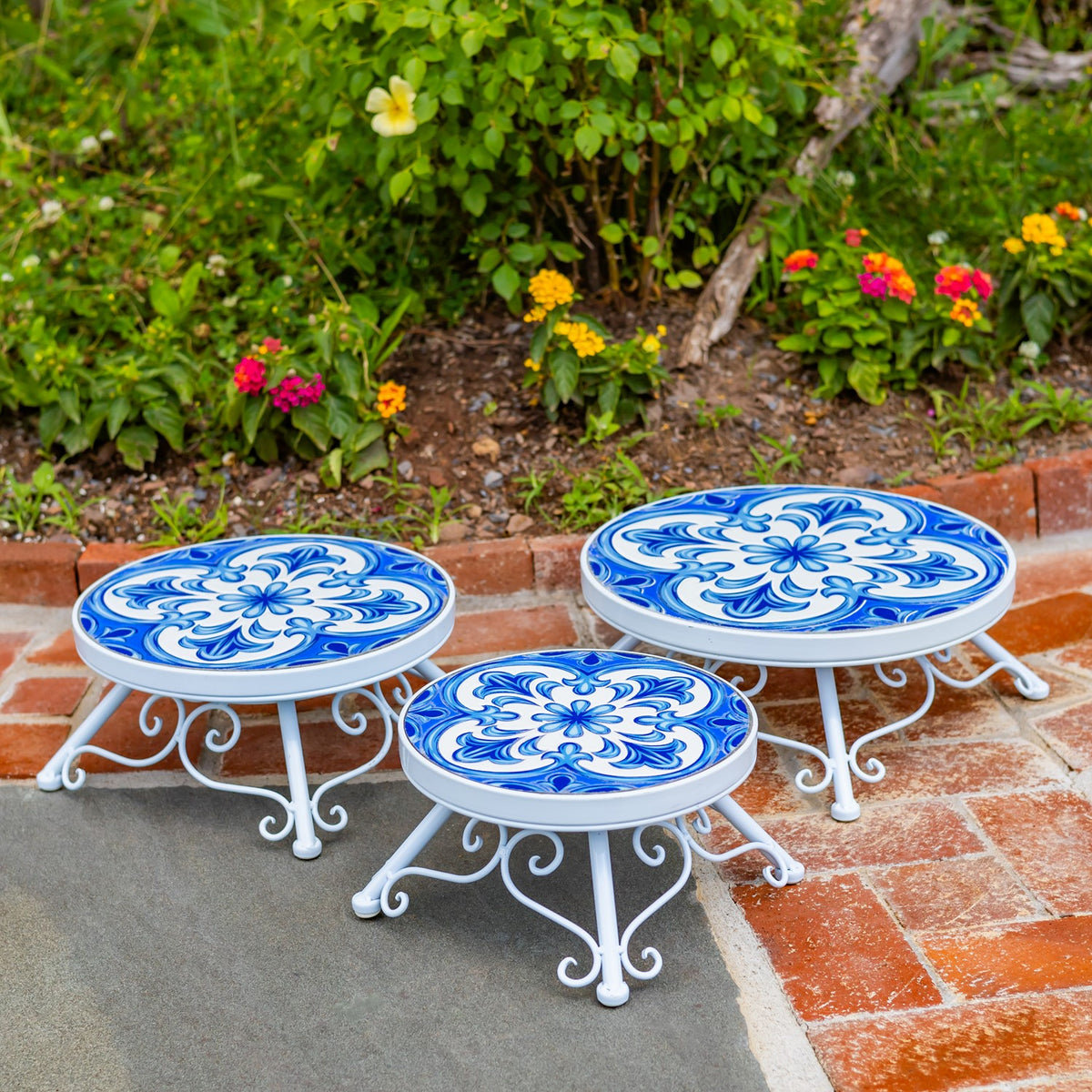 Three empty white metal plant stands with round blue floral mosaic tops arranged on a brick patio.