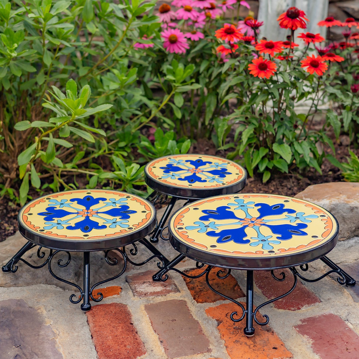 Three round mosaic plant stands in different heights arranged among flowers on a brick path.