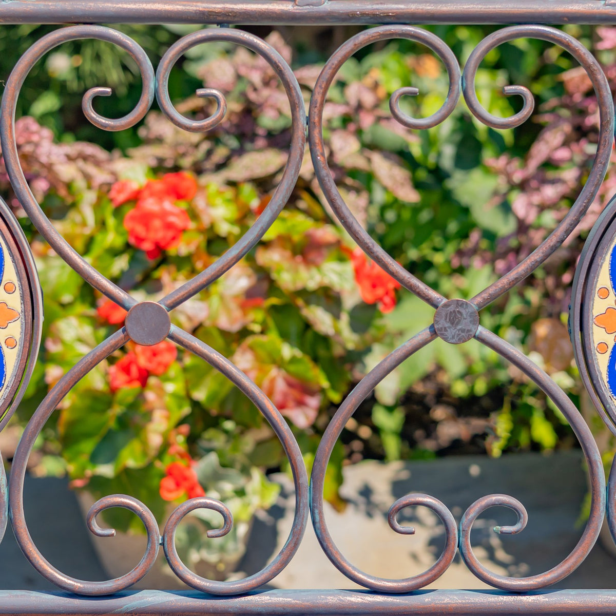 Close-up of curled bronze-colored metal scrollwork with garden flowers softly visible behind it.