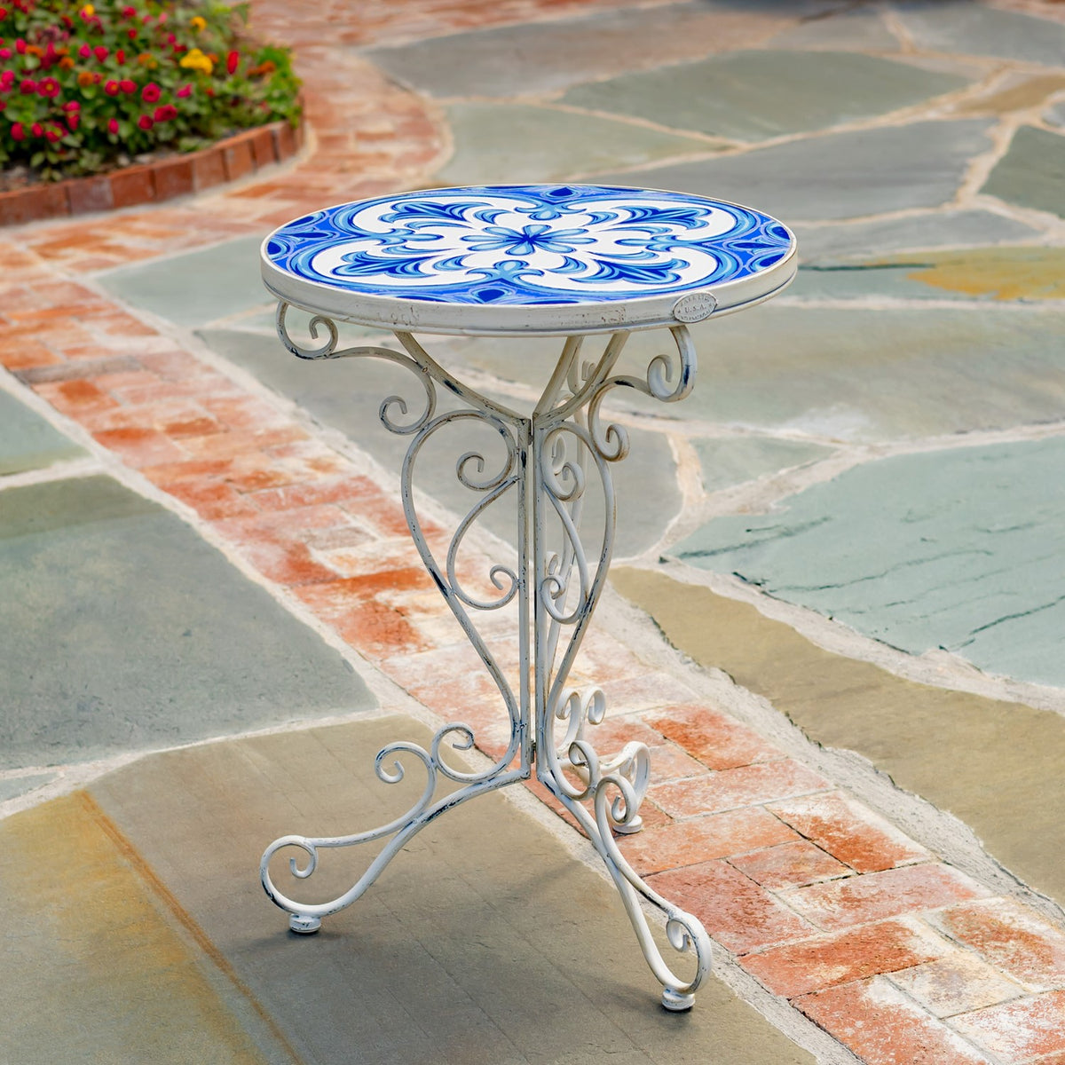 A round blue-and-white mosaic plant stand on a curled silver-tone metal base, displayed on a stone patio.